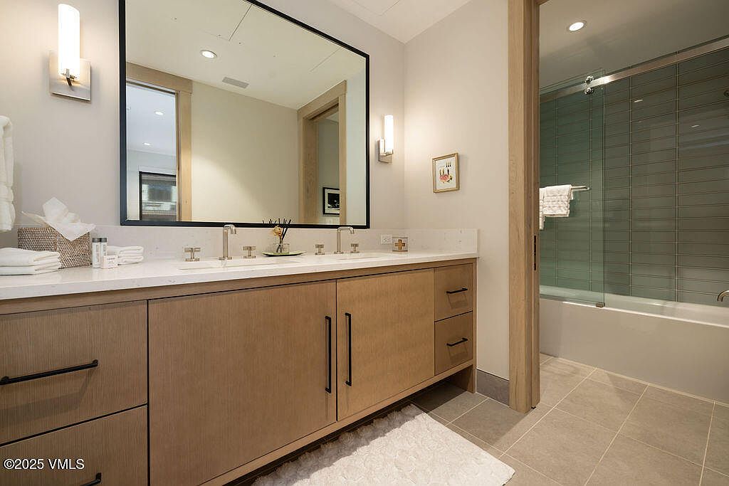 This is a well-lit bathroom featuring a double vanity with a white countertop and light wood cabinetry. A large mirror with a black frame hangs above the vanity, reflecting the interior of another room. The bathroom also includes a bathtub with a glass shower door and tiled walls, creating a clean and modern aesthetic.