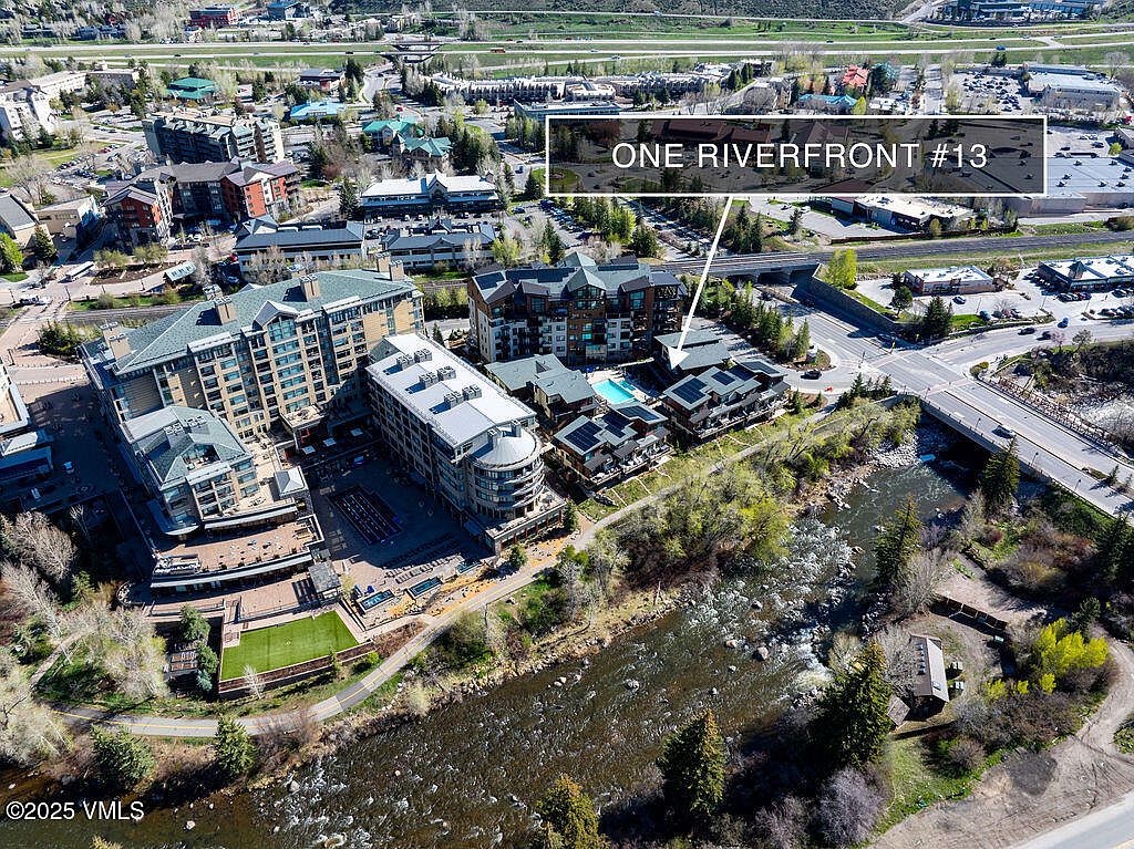 This aerial view showcases a luxurious condominium complex situated alongside a river. The buildings feature modern architecture with a mix of materials, including stone and wood accents. A pool and outdoor amenities are visible, enhancing the property's appeal, while the surrounding landscape includes lush greenery and a bridge, adding to the scenic environment.