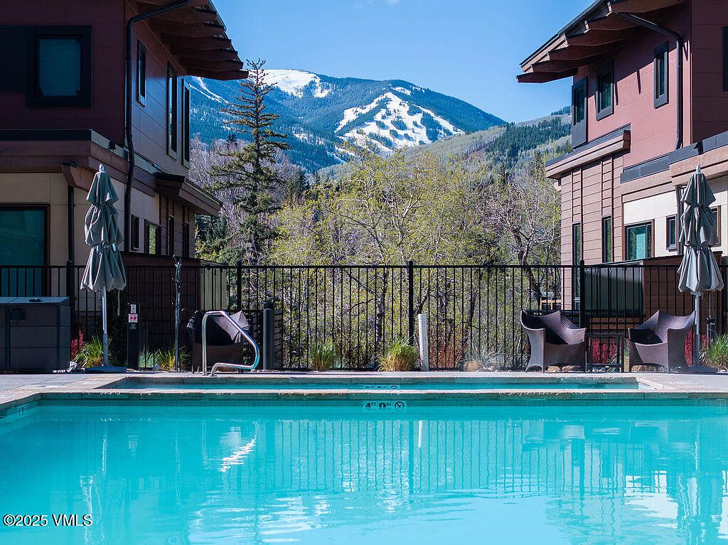 This image showcases a serene pool area nestled between two modern residential buildings, with a stunning mountain view in the background. The clear blue water of the pool reflects the sky, while lounge chairs sit poolside, inviting relaxation. A black metal fence provides a subtle boundary, and the overall scene exudes a luxurious and tranquil atmosphere.