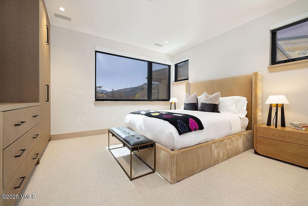 This is a well-lit primary bedroom featuring a large window with mountain views. The room includes a king-size bed with a neutral-toned upholstered headboard and a decorative throw. A wooden nightstand with a modern lamp and a bench at the foot of the bed add to the room's sophisticated and comfortable ambiance.