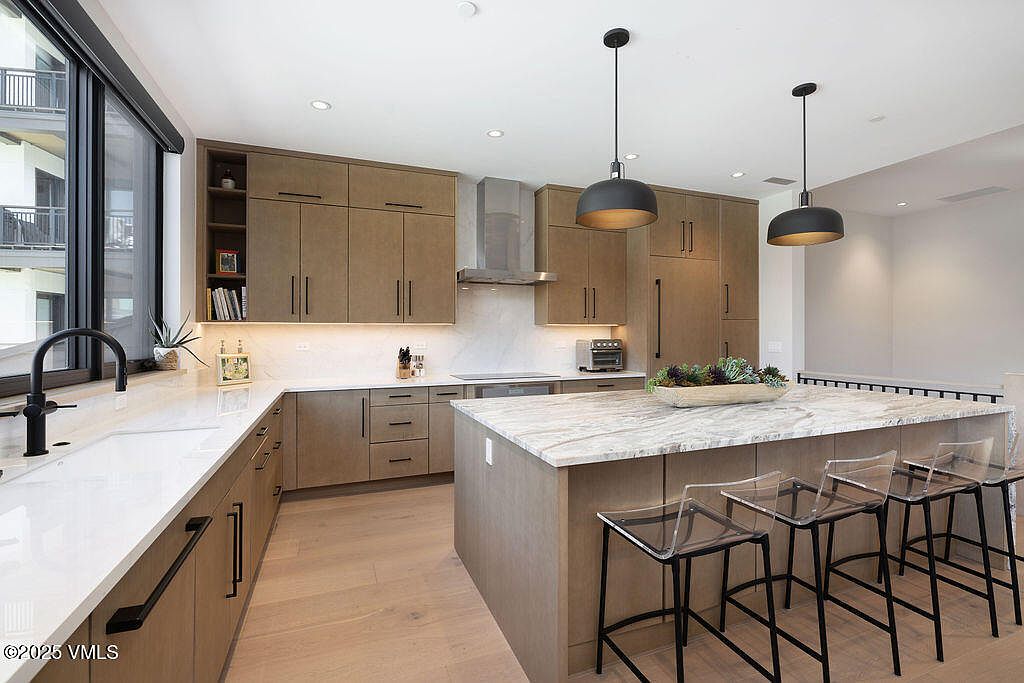 This is a well-lit, modern kitchen featuring sleek, light brown cabinetry with black hardware and a large island with a marble countertop. The kitchen is equipped with stainless steel appliances and illuminated by pendant lighting. The perspective is from a wide angle, showcasing the entire kitchen layout and its connection to the adjacent living space.