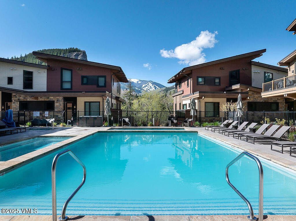 This image showcases a pristine outdoor pool area, flanked by modern residential buildings with a mountain backdrop. The pool's clear blue water invites relaxation, while lounge chairs offer a comfortable space for sunbathing. The setting suggests a luxurious and well-maintained community amenity.
