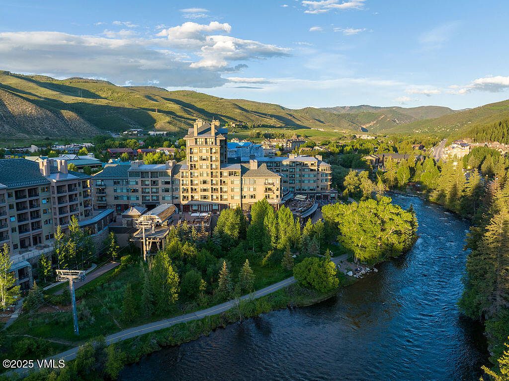 This aerial view showcases a luxurious multi-story building complex nestled amidst lush greenery and a flowing river. The architecture features a blend of modern and classic elements, with multiple balconies and terraces offering scenic views. The surrounding landscape includes rolling hills and mature trees, creating a serene and upscale atmosphere.