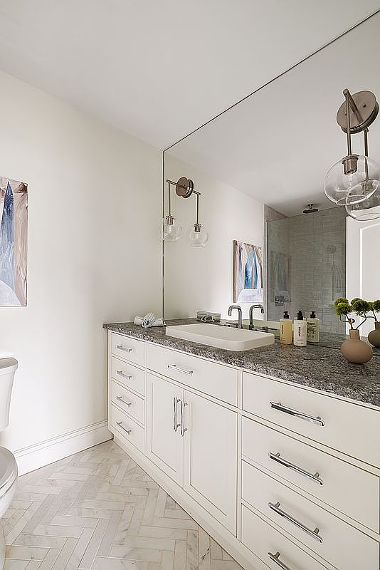 This bright and clean bathroom features a white vanity with a dark granite countertop and a rectangular vessel sink. The space is accented by elegant wall-mounted light fixtures, a large mirror, and a herringbone-patterned marble floor. The overall aesthetic is modern and sophisticated, offering a polished look suitable for a guest or secondary bathroom.