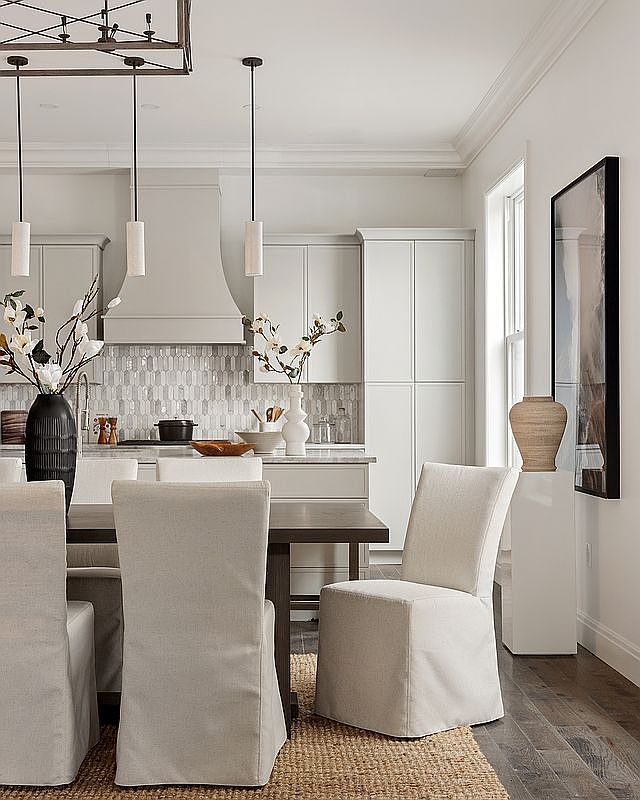 This elegant, open-concept kitchen and dining area features a sophisticated neutral palette with light gray cabinetry, a custom range hood, and a textured mosaic tile backsplash. A dark wood dining table is surrounded by slipcovered chairs, illuminated by modern pendant lighting, creating a warm and inviting atmosphere. The space is accented with tasteful decor, including floral arrangements and a large framed piece of art, emphasizing a high-end, transitional design aesthetic.