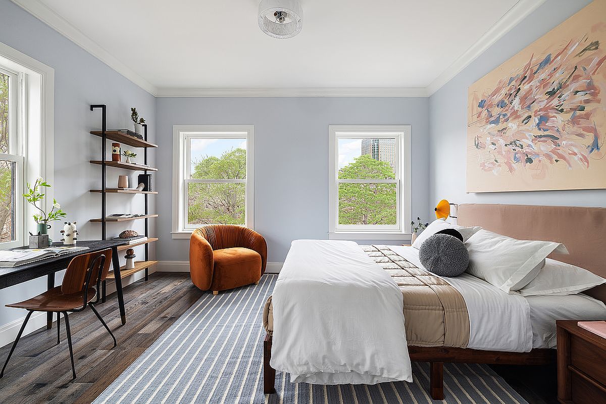 This bright and airy primary bedroom features a contemporary design with light blue walls, dark wood flooring, and a large abstract painting above the bed. The room is furnished with a comfortable bed, a stylish burnt-orange accent chair, and a sleek black shelving unit that doubles as a workspace. Large windows provide ample natural light and a pleasant view of the surrounding trees, creating a serene and inviting atmosphere.