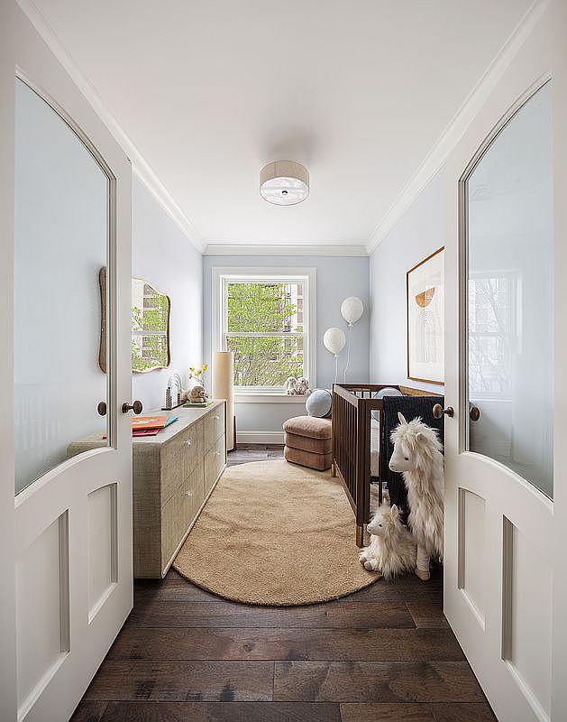 This charming nursery is captured from a doorway perspective, showcasing a warm and inviting atmosphere with soft blue walls and rich dark wood flooring. The room features a wooden crib, a plush beige armchair, a textured oval rug, and a long dresser, all accented by whimsical stuffed animals and soft lighting. The space feels cozy and well-curated, perfect for a nursery or small child's bedroom.