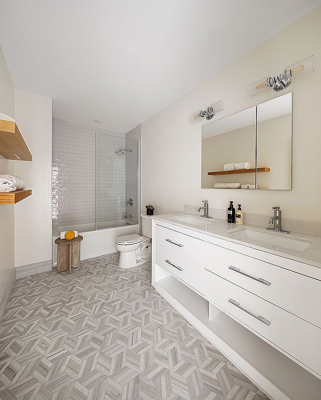 This modern, bright bathroom features a double vanity with white cabinetry, a sleek quartz countertop, and a large mirror with integrated lighting. The space includes a tiled shower-tub combination with a glass enclosure and a distinctive geometric patterned floor, creating a clean and contemporary aesthetic. The perspective is a wide-angle shot from the doorway, showcasing the efficient layout and high-end finishes.