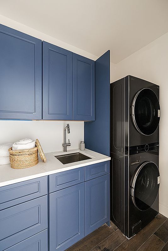 This modern laundry room features striking navy blue cabinetry paired with a clean white countertop and an integrated sink. A sleek, stacked black washer and dryer unit stands beside the cabinets, creating a sophisticated and functional workspace. The dark wood flooring adds warmth to the space, which is illuminated by bright, neutral lighting.