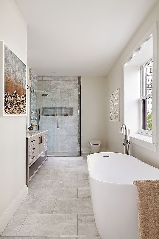 This elegant primary bathroom features a sleek freestanding soaking tub, a spacious glass-enclosed walk-in shower with marble-style tiling, and a modern vanity with ample storage. The room is bathed in natural light from a large window, creating a bright, serene, and luxurious atmosphere. The neutral color palette and clean lines emphasize a sophisticated, contemporary design.