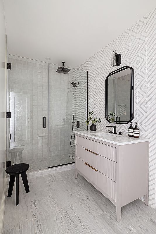 This modern primary bathroom features a sleek vanity with a marble countertop, a black-framed mirror, and a walk-in glass shower with a rainfall showerhead. The space is accented by geometric patterned wall tiles and light wood-look flooring, creating a clean, contemporary aesthetic. A small black stool in the foreground adds a functional and stylish touch to the well-lit, minimalist design.