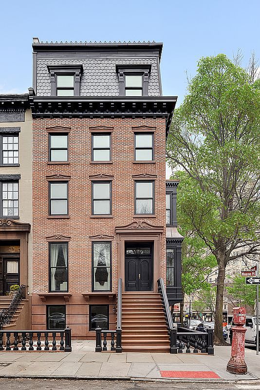 This classic brownstone townhouse features a striking red brick facade with a prominent mansard roof and decorative dormer windows. A grand set of stone steps leads up to a stately double-door entrance, framed by elegant architectural detailing. The perspective is a straight-on street-level shot, capturing the historic charm of the building alongside a mature tree and a vintage-style fire hydrant.