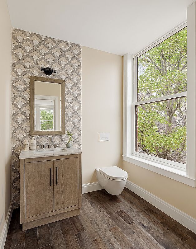 This elegant powder room features a sophisticated vanity with a marble countertop, set against a striking patterned wallpaper accent wall. A wall-mounted toilet and rich, dark wood-toned flooring create a modern and clean aesthetic, while a large window provides ample natural light and a serene view of lush green trees. The perspective is a straight-on shot that highlights the balance between the textured wallpaper and the minimalist fixtures.