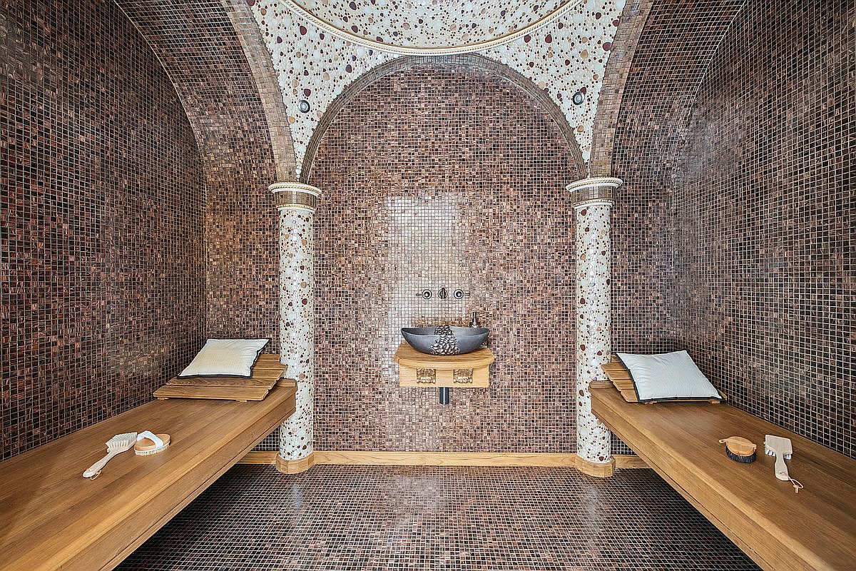 This luxurious spa-inspired steam room features intricate brown mosaic tile work throughout the walls and floor, accented by two decorative columns with a speckled stone finish. Two wooden benches with plush pillows provide seating on either side, while a central stone vessel sink mounted on a wooden shelf serves as a focal point. The arched ceiling and warm lighting create an intimate, high-end atmosphere perfect for relaxation.
