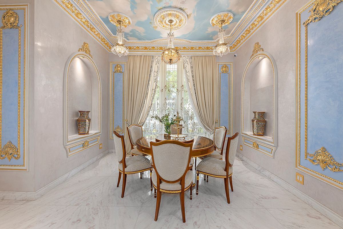 This opulent dining room features a round glass-top table surrounded by four elegant, upholstered chairs, set against a backdrop of neoclassical design. The space is defined by intricate gold-leaf crown molding, a hand-painted sky-themed ceiling with three crystal chandeliers, and symmetrical arched wall niches housing decorative vases. The marble flooring and soft, draped curtains enhance the luxurious, formal atmosphere of this sophisticated dining area.