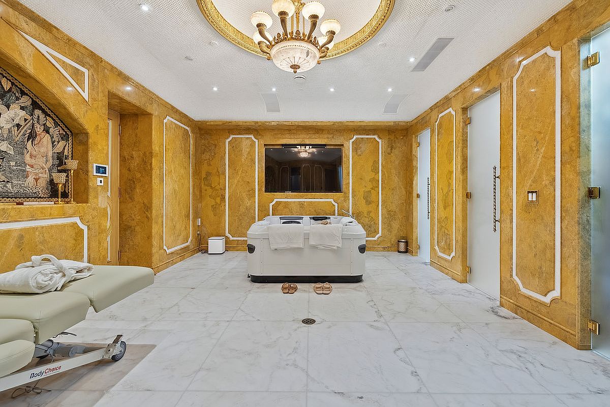 This opulent spa-inspired bathroom features striking gold-toned marble walls with white trim, a central white jetted tub, and a massage table in the foreground. The room is illuminated by a grand crystal chandelier centered under a decorative ceiling medallion, creating a luxurious and serene atmosphere. The perspective is a wide, symmetrical shot that emphasizes the high-end finishes and the spacious, tranquil layout of the wellness area.