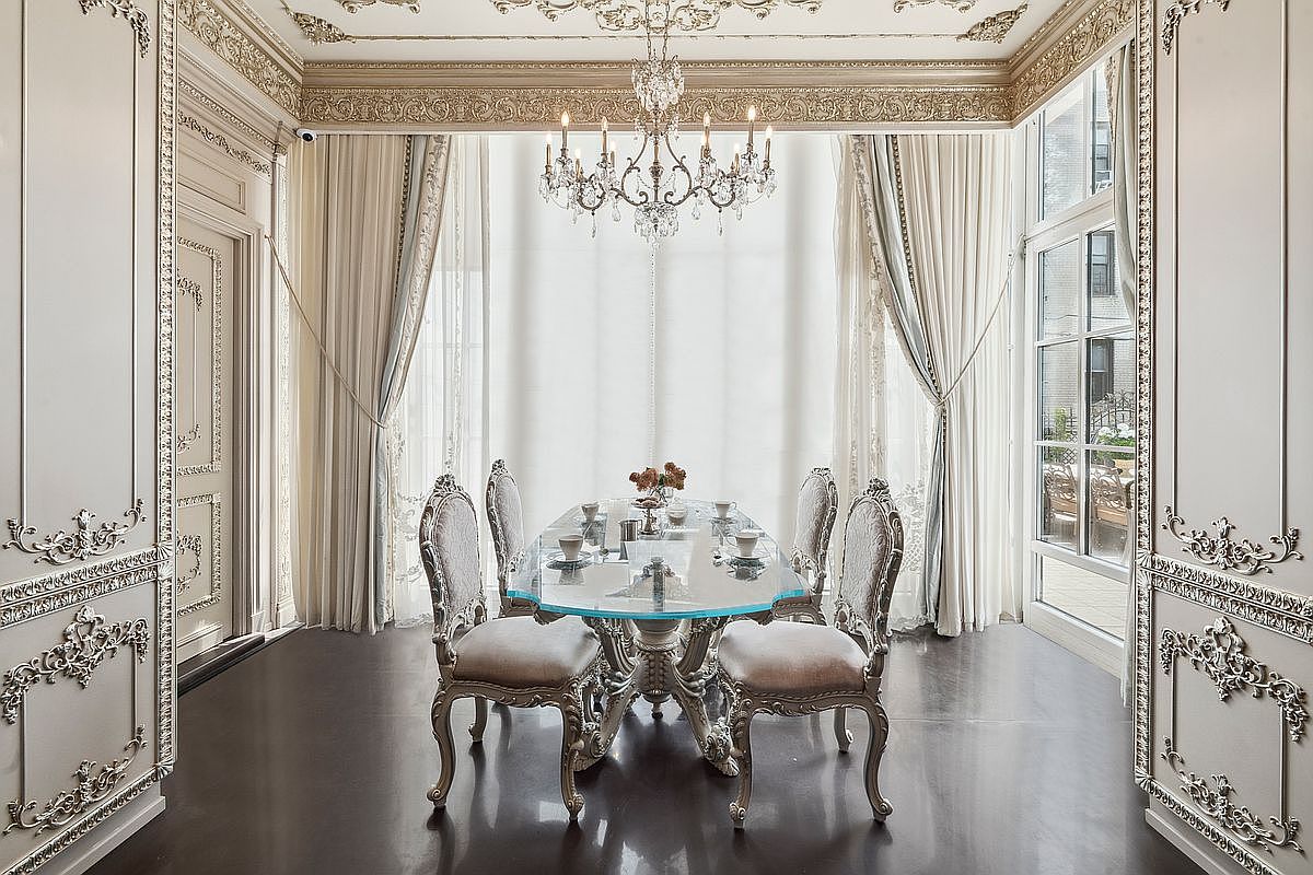 This elegant dining room features a sophisticated neoclassical design with ornate wall moldings, a crystal chandelier, and floor-to-ceiling drapery. A glass-topped table with intricately carved chairs sits at the center, resting on dark hardwood floors that contrast beautifully with the light, neutral color palette. The room is bathed in natural light from large windows, creating a luxurious and formal atmosphere perfect for refined entertaining.