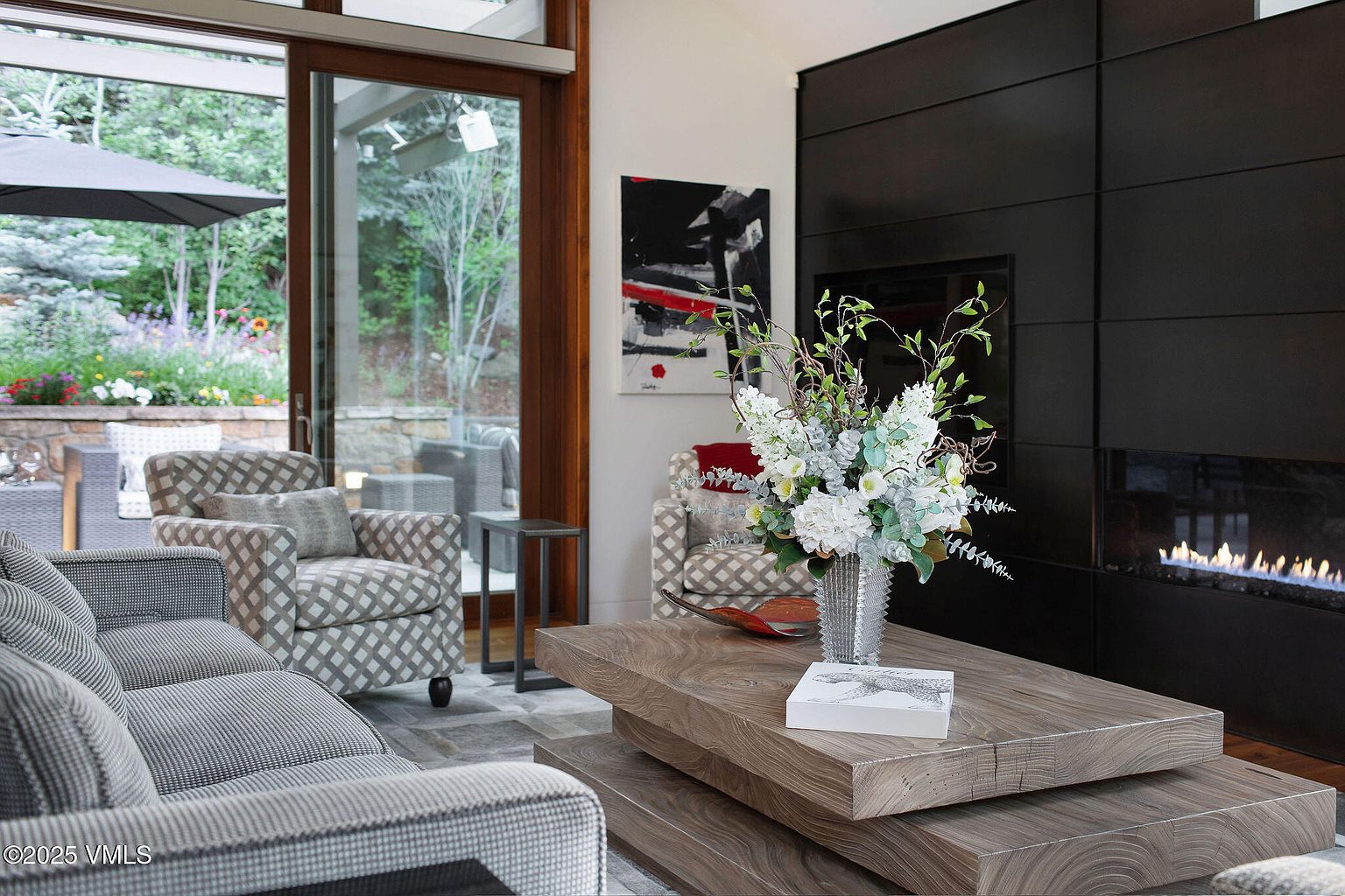 This is an interior shot of a living room featuring a modern fireplace with a dark surround, a layered wooden coffee table with floral arrangement, and comfortable seating including a patterned armchair and a gray sofa. Large sliding glass doors offer a view of an outdoor patio and garden, creating a seamless indoor-outdoor living experience. The perspective is from a low angle, emphasizing the depth and spaciousness of the room.