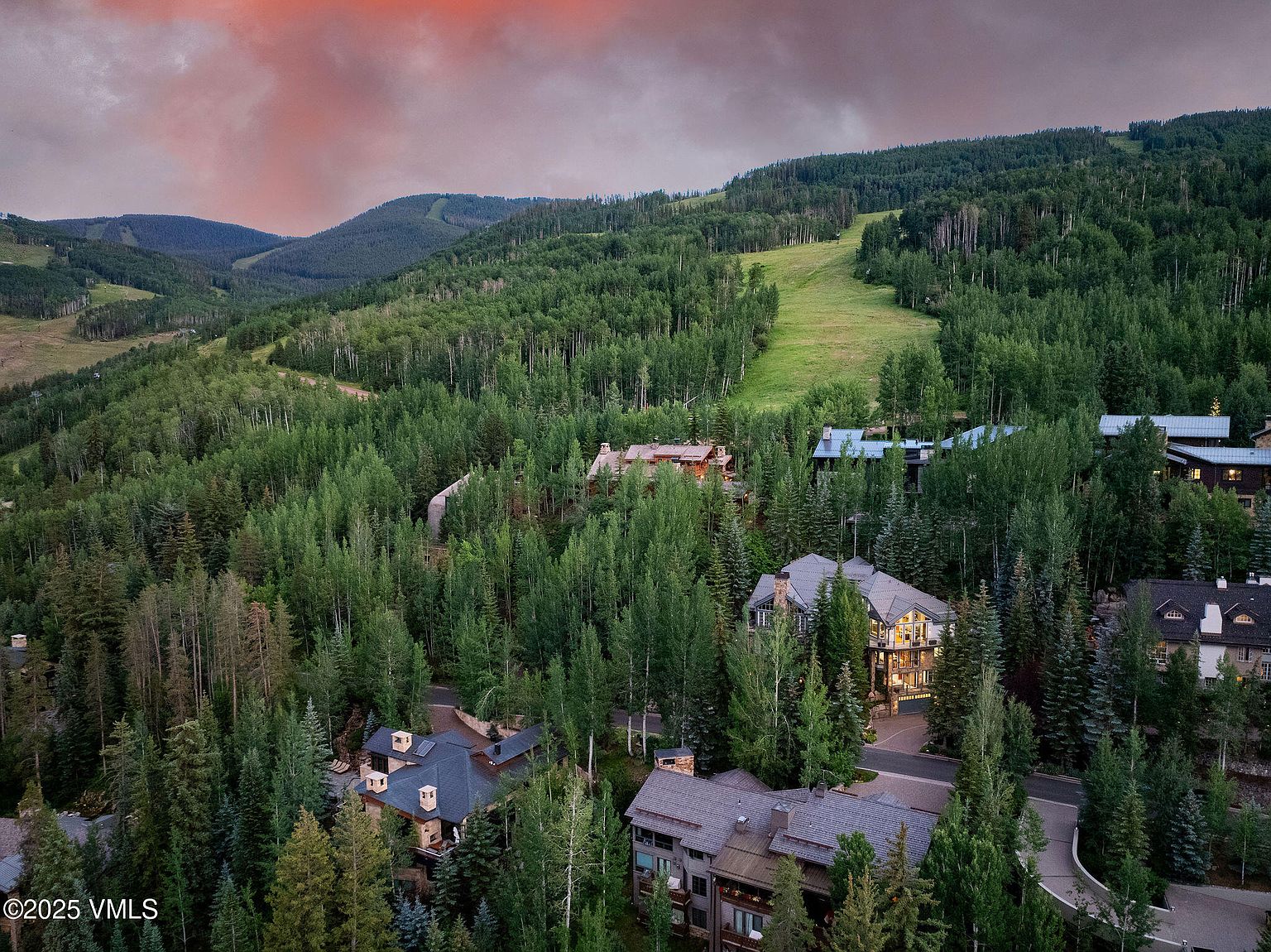 This aerial view showcases a collection of luxury homes nestled amidst a dense forest landscape, with a ski slope visible in the background. The homes feature varying architectural styles, with prominent rooflines and well-maintained exteriors, suggesting a high-end residential community. The surrounding greenery and the distant mountains create a serene and exclusive atmosphere.