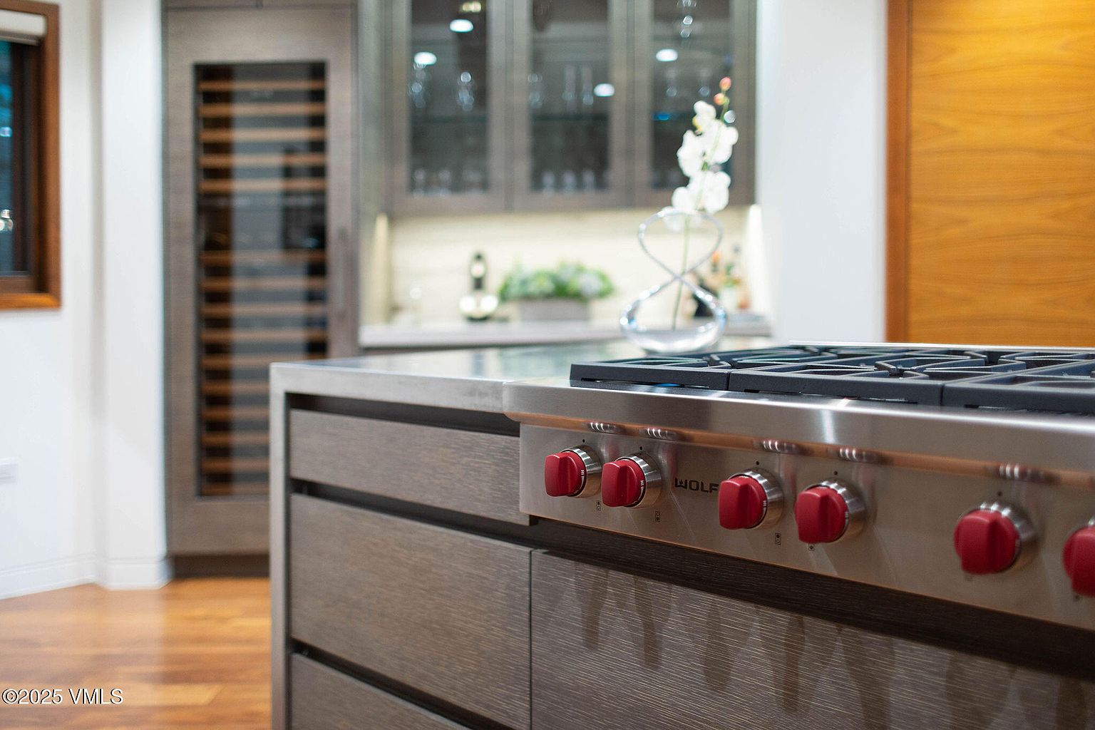 This image showcases a modern kitchen with a focus on the Wolf range, featuring red knobs and a stainless steel finish. The cabinetry is a sleek, dark wood, complemented by a light countertop. In the background, there's a wine cooler and glass-front cabinets, adding to the upscale feel of the space.