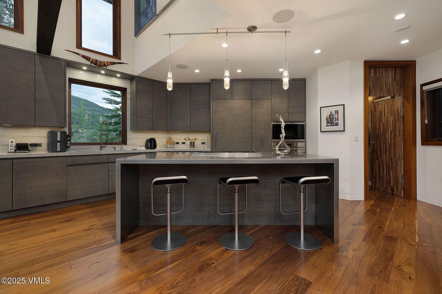This is a modern kitchen featuring dark wood cabinetry and a large island with seating. Stainless steel appliances are integrated seamlessly into the design. Three pendant lights hang above the island, and hardwood floors add warmth to the space. A window provides natural light and a view of the outdoors.