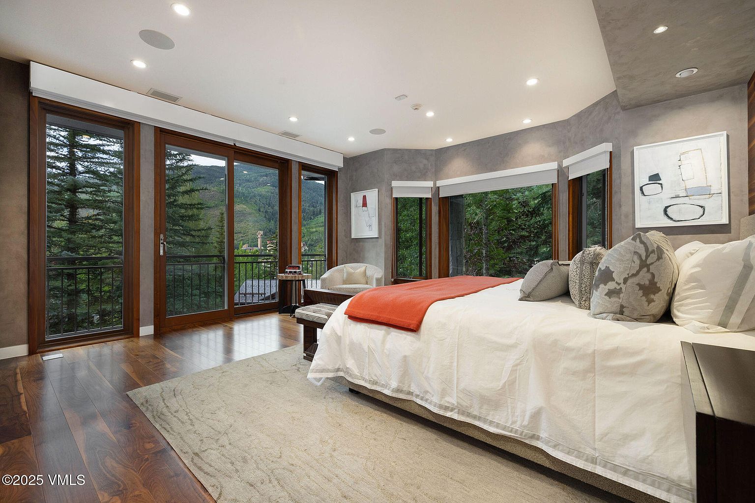 This is a luxurious primary bedroom featuring hardwood floors, a large bed with white linens and an orange throw, and floor-to-ceiling windows offering views of the surrounding landscape. The room is decorated in neutral tones with gray accent walls and modern artwork, creating a serene and sophisticated atmosphere. A balcony is accessible through sliding glass doors.