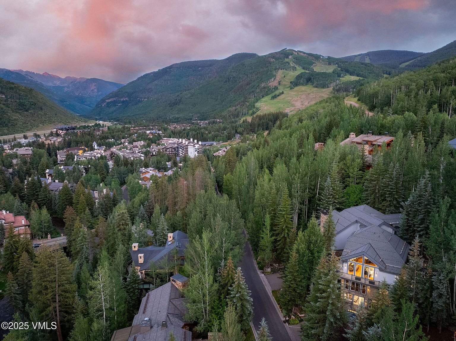 This aerial view showcases a luxurious property nestled among lush green trees, with a charming town visible in the distance. The home features a dark roof and multiple stories, suggesting ample living space. The surrounding landscape includes mountains and a picturesque sky, creating a serene and desirable setting.