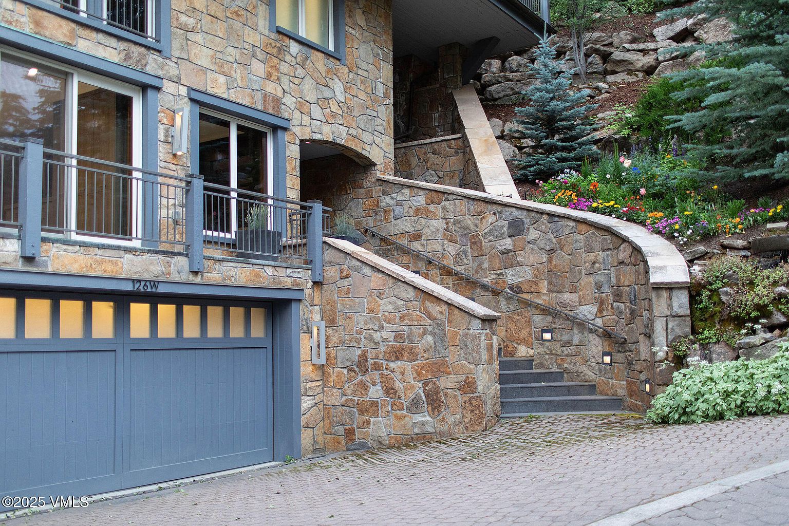 This is a front exterior view of a stone-clad house featuring a garage, a stone staircase leading to the entrance, and a small balcony. The house is built into a hillside with landscaping and flowers visible. The overall impression is one of quality construction and integration with the natural surroundings.