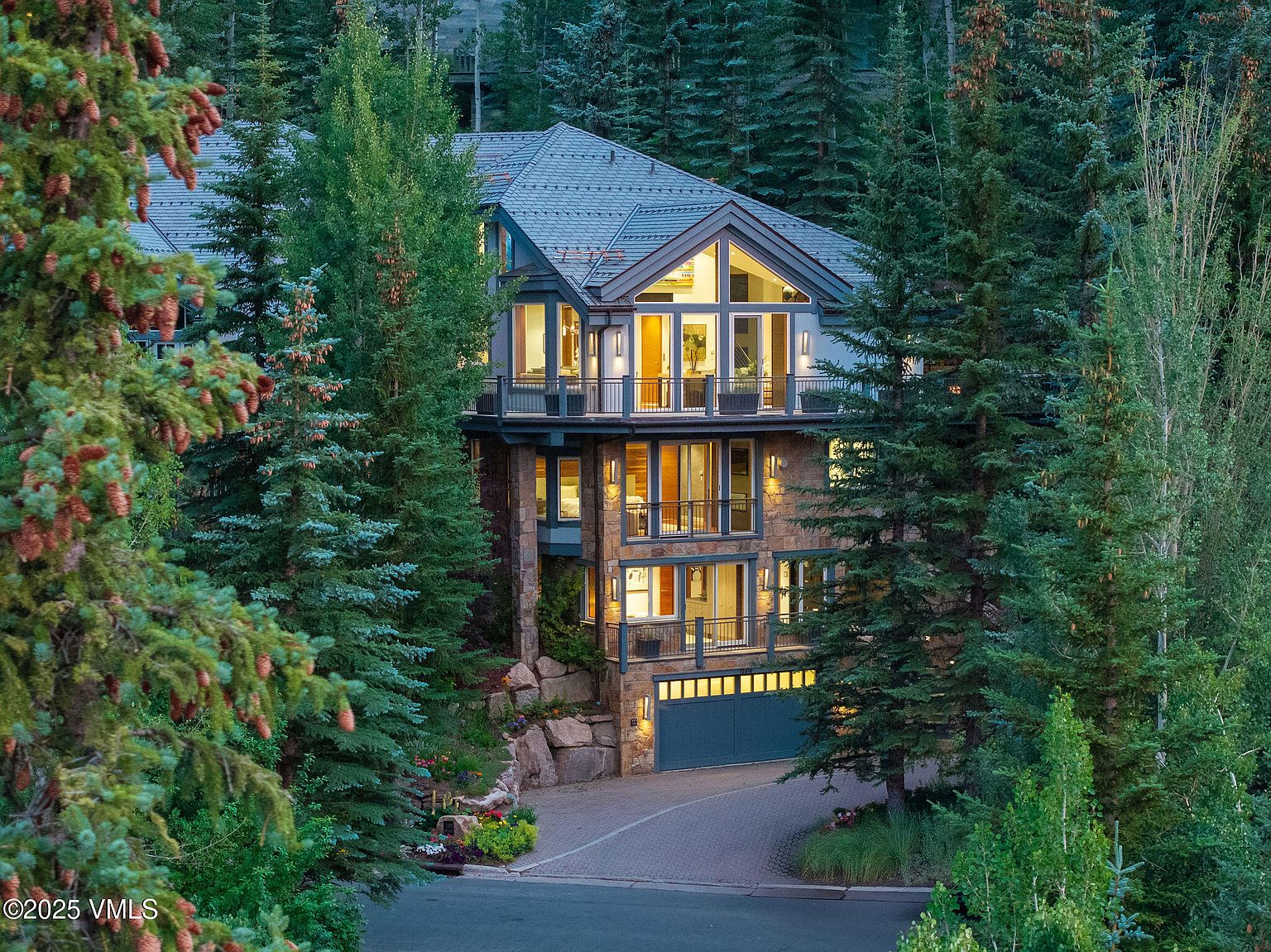 This is a front exterior view of a multi-story luxury home nestled among tall evergreen trees. The house features a stone facade, multiple balconies with railings, and large windows that allow ample natural light. A paved driveway leads to a garage, and the landscaping includes mature trees and shrubs, creating a sense of privacy and seclusion.