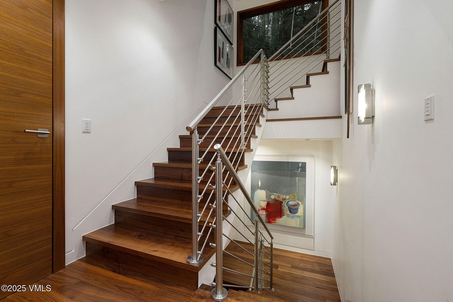 This interior shot showcases a modern staircase with wooden steps and a sleek stainless steel railing. The walls are painted white, and artwork is visible in the background, adding a touch of sophistication. The hardwood flooring extends into the hallway, creating a seamless transition.
