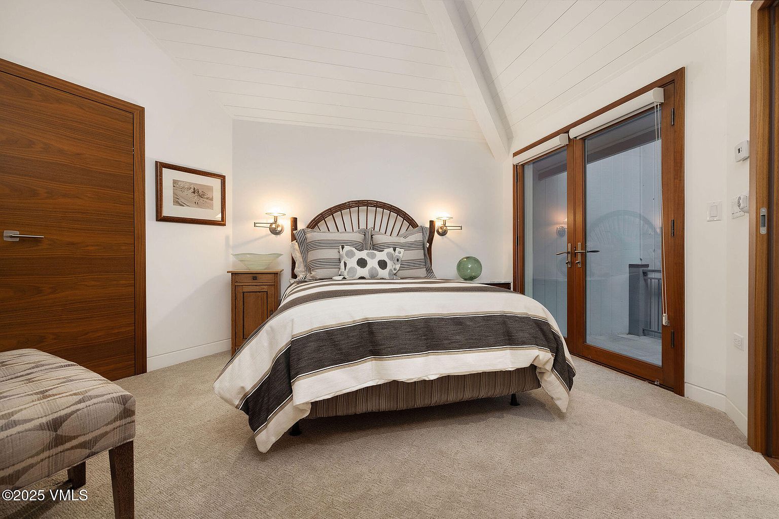 This is a cozy bedroom featuring a bed with striped bedding and a wooden headboard. The room has white walls and a vaulted ceiling with wooden beams, creating a bright and airy atmosphere. There are wooden doors and trim, along with bedside tables and lamps, adding warmth and character to the space.
