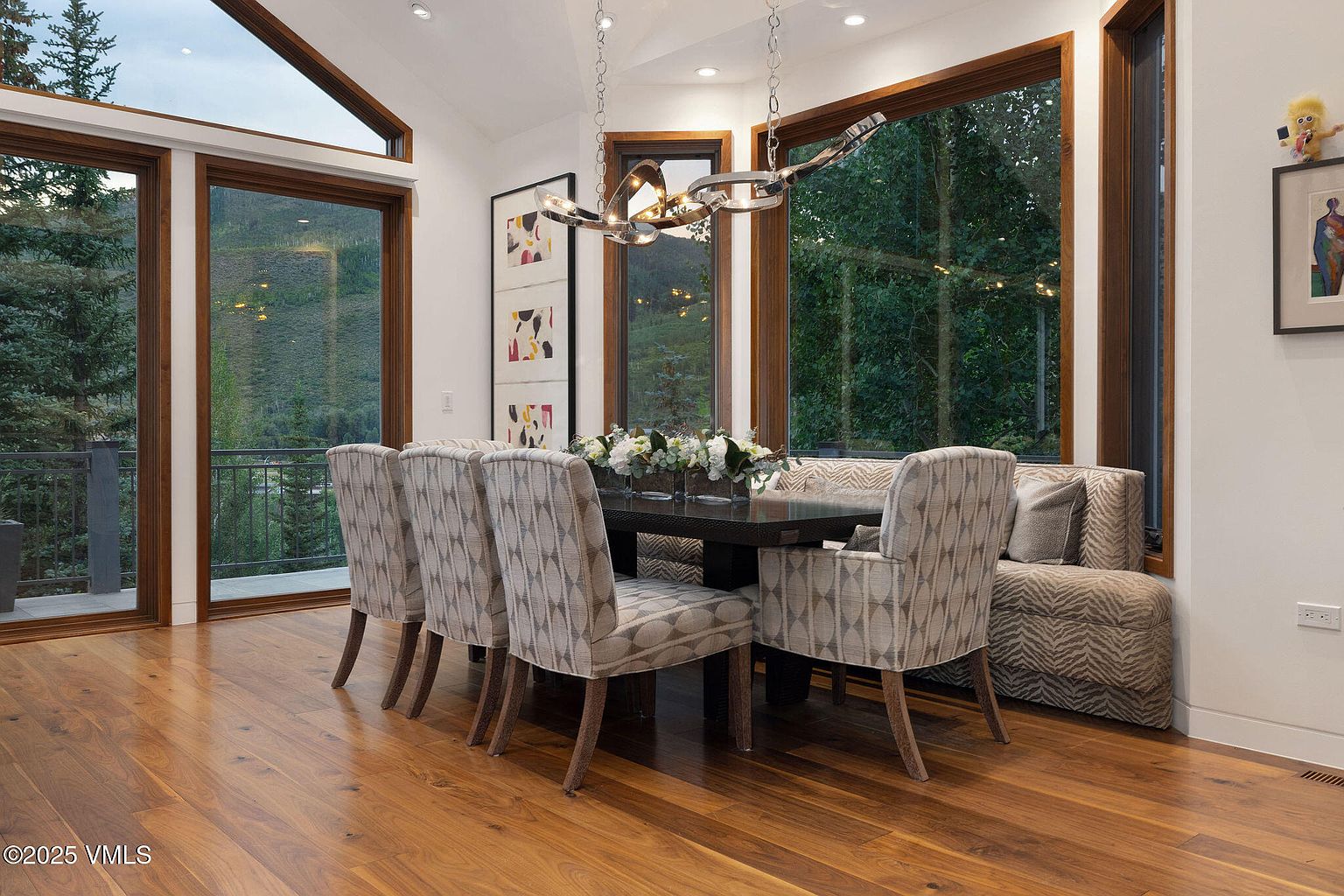 This is a dining room featuring a dark wood table set with flowers, surrounded by upholstered chairs with a geometric pattern and a built-in bench. Large windows offer a view of the surrounding landscape, and a modern chandelier hangs above the table. The room has a sophisticated and inviting atmosphere.
