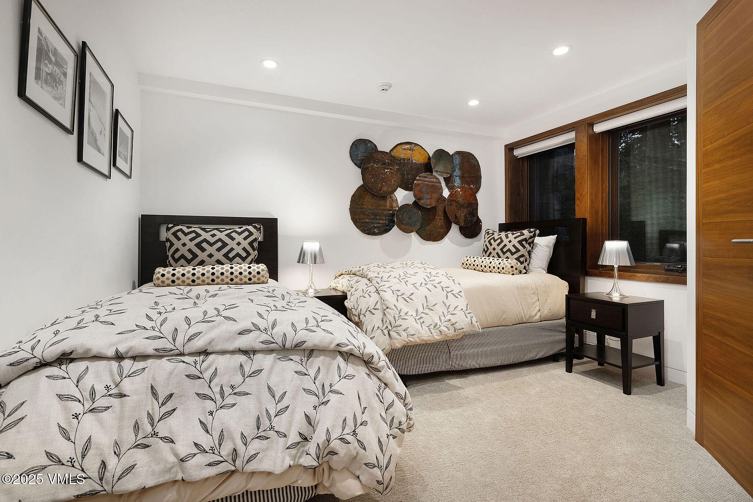 This is a guest bedroom featuring two twin beds with patterned bedding and decorative pillows. The room has a neutral color palette with white walls and beige carpeting, complemented by dark wood headboards and side tables. A unique circular metal wall art piece adds visual interest above one of the beds, and natural light filters through the windows, creating a cozy and inviting atmosphere.
