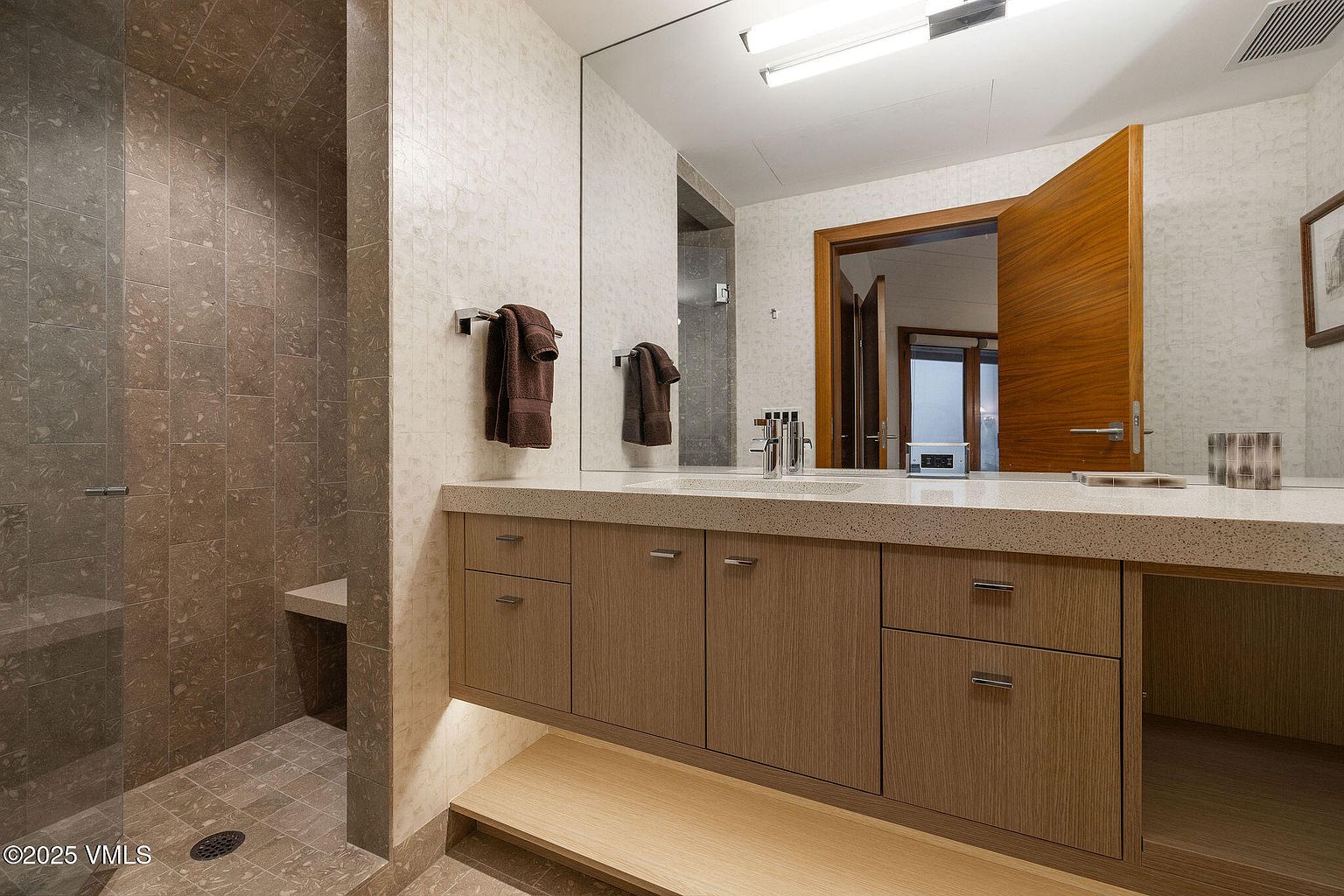 This is a well-lit primary bathroom featuring a modern vanity with wood-grain cabinets and a light-colored countertop. A large mirror reflects the open doorway and a glimpse of another room, enhancing the sense of space. The shower area is tiled in a darker shade, providing a contrast to the lighter tones of the rest of the bathroom.