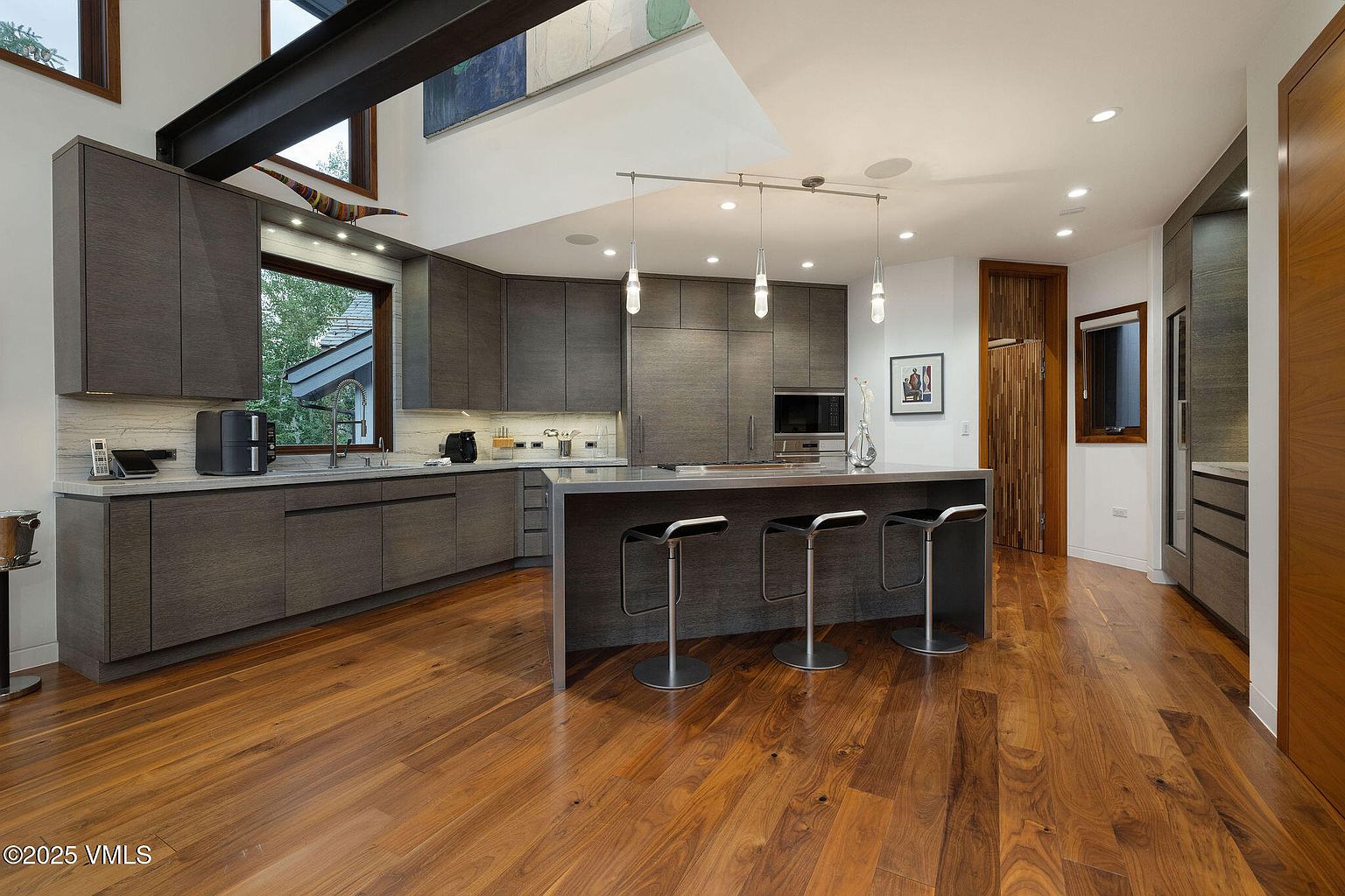This is a modern kitchen with dark wood cabinetry and stainless steel appliances. A large island with bar seating anchors the space, illuminated by pendant lighting. The hardwood floors add warmth, and the overall design is sleek and sophisticated.
