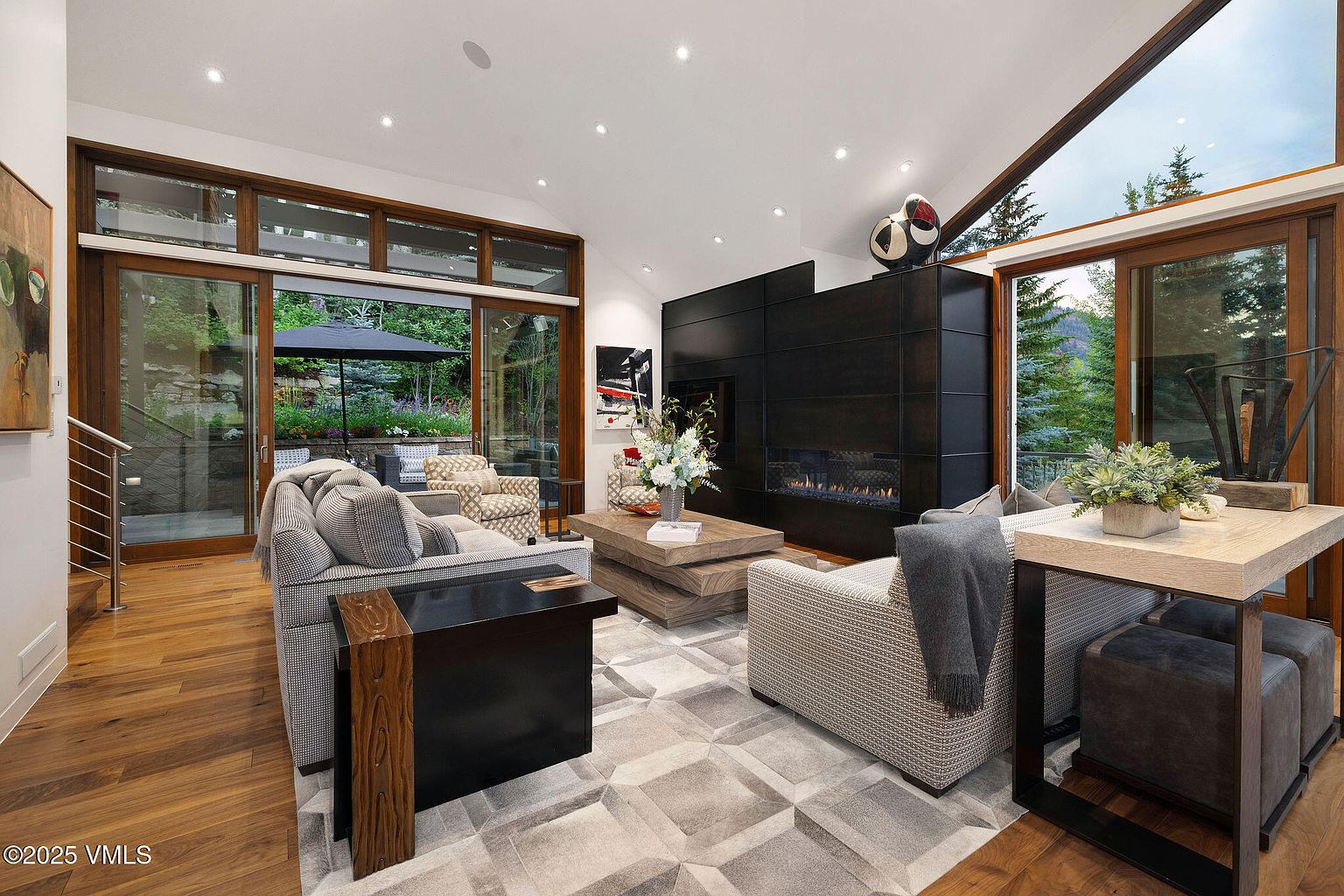 This is a well-lit living room featuring a modern fireplace, comfortable seating, and large windows that offer a view of the outdoors. The room is decorated with neutral colors and has a contemporary style. The open floor plan and natural light create a welcoming and spacious atmosphere.