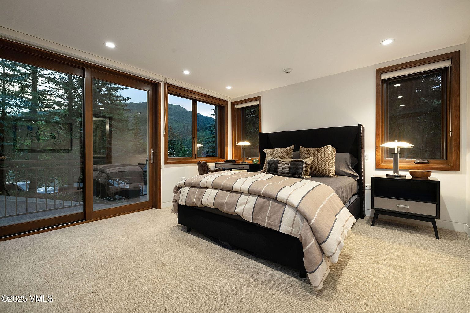 This primary bedroom features a large bed with a dark headboard and a striped comforter, flanked by a modern nightstand with a lamp. Large windows offer views of the surrounding landscape, bringing natural light into the room. The room is carpeted and has a neutral color palette, creating a serene and inviting atmosphere.