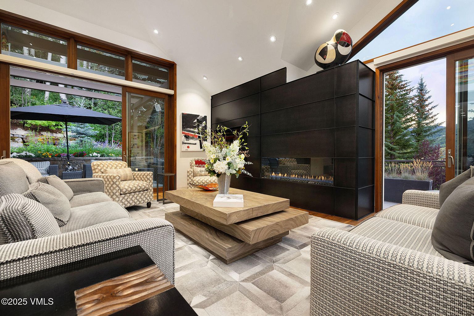 This is an interior shot of a luxurious living room featuring a modern fireplace with a dark paneled surround. The room is furnished with comfortable sofas and a unique layered wooden coffee table, set upon a geometric patterned rug. Large windows offer views of the outdoor landscape, enhancing the room's connection to nature.