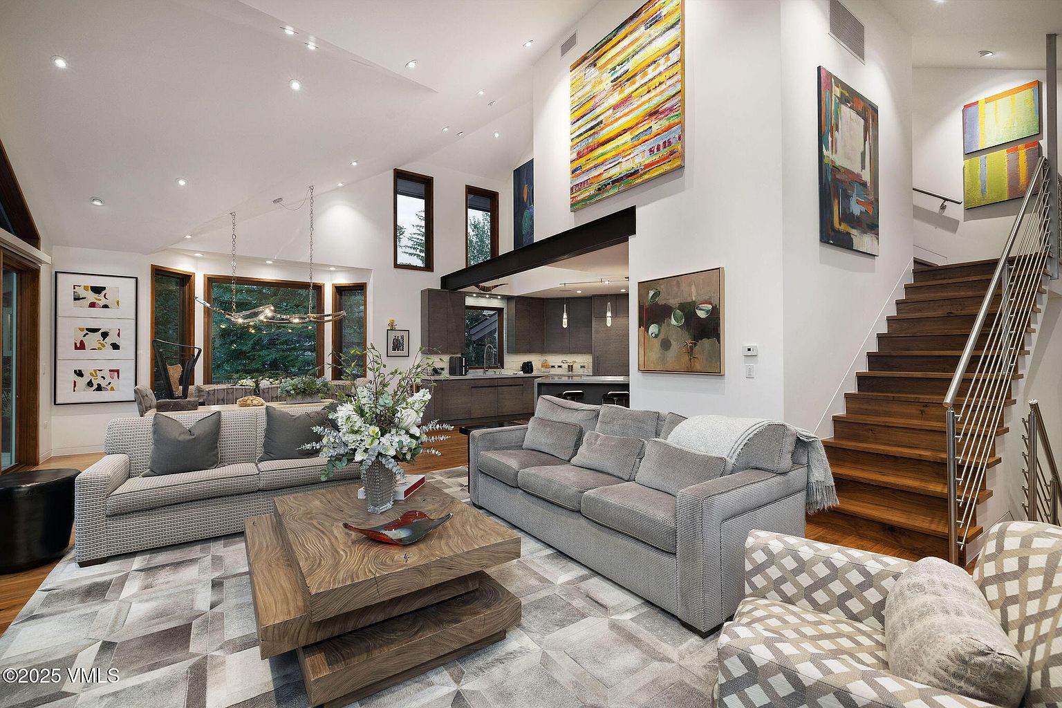 This is an interior shot of a modern living room featuring two sofas, a unique layered coffee table, and an armchair. The room is decorated with contemporary art and has a staircase leading to the upper level. The overall impression is one of sophisticated comfort and style.