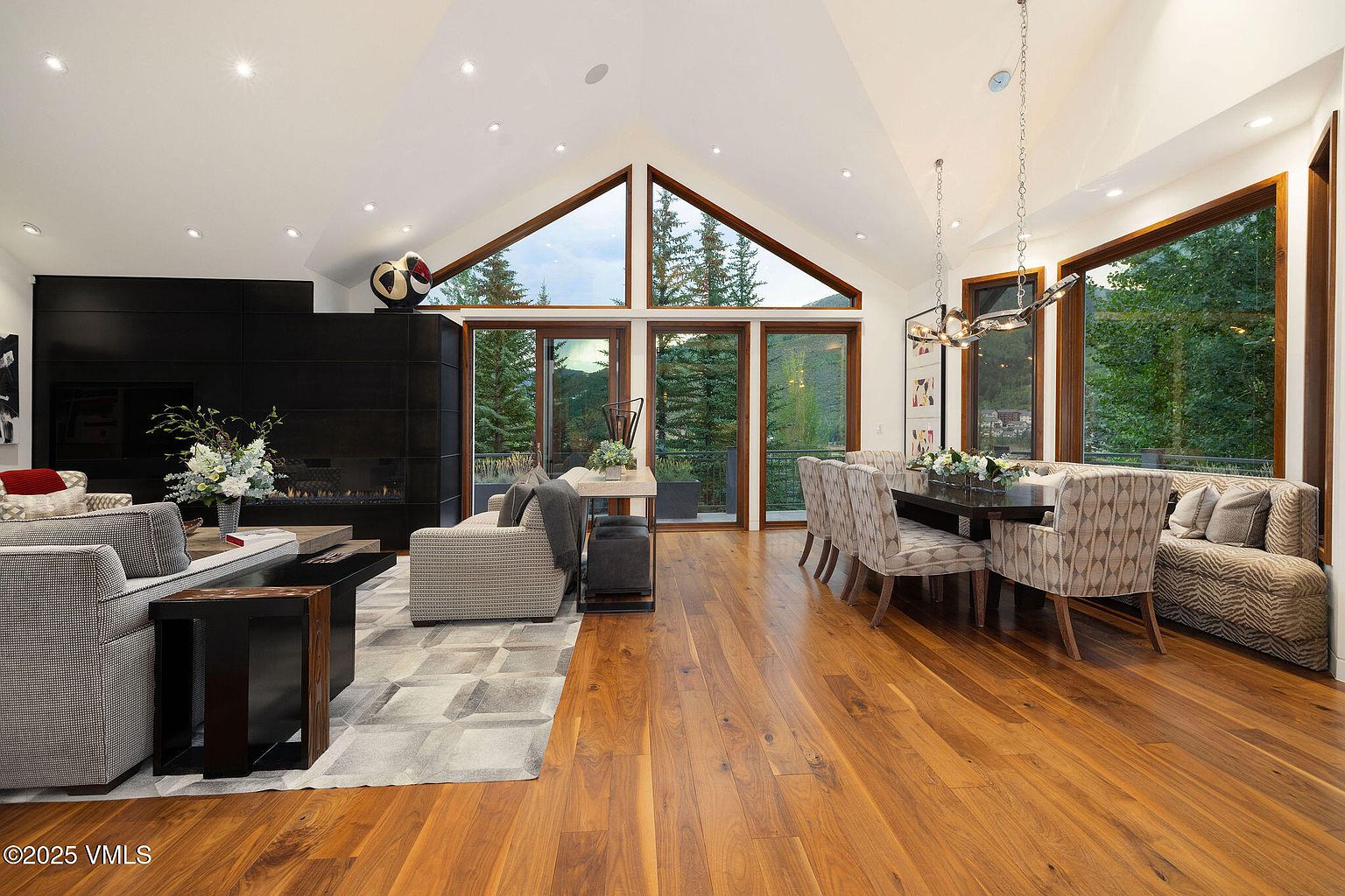 This is an interior shot of a luxurious living and dining area featuring hardwood floors, high vaulted ceilings, and large windows offering views of the surrounding landscape. The living area includes comfortable seating around a modern fireplace, while the dining area features an elegant table and chairs. The overall impression is one of spaciousness, sophistication, and connection to nature.
