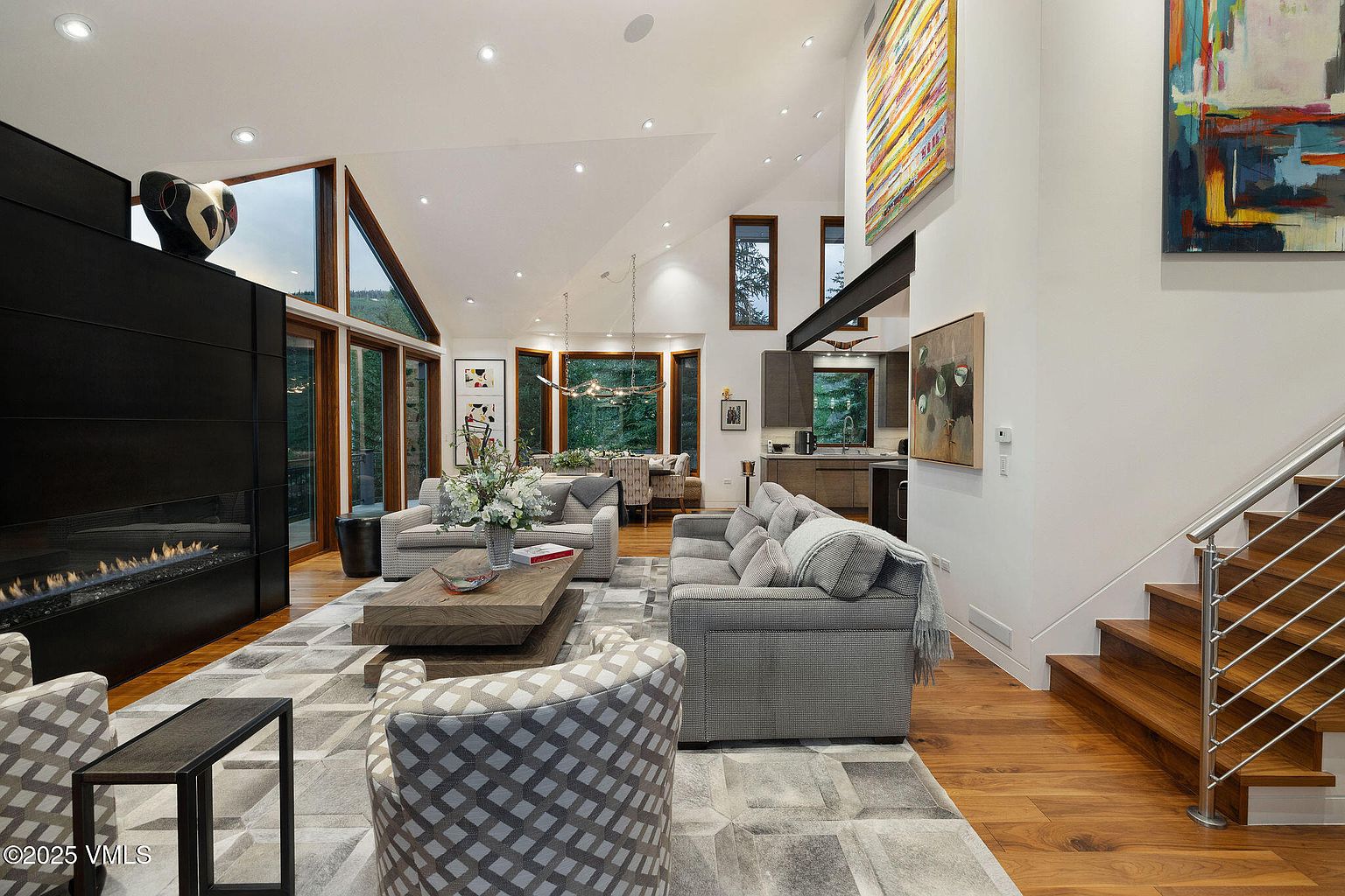 This is a spacious living room featuring a modern fireplace, comfortable seating arrangements with sofas and armchairs, and a stylish coffee table. Large windows provide ample natural light and views of the surrounding landscape. The room is decorated with contemporary art and has hardwood floors, creating a warm and inviting atmosphere.