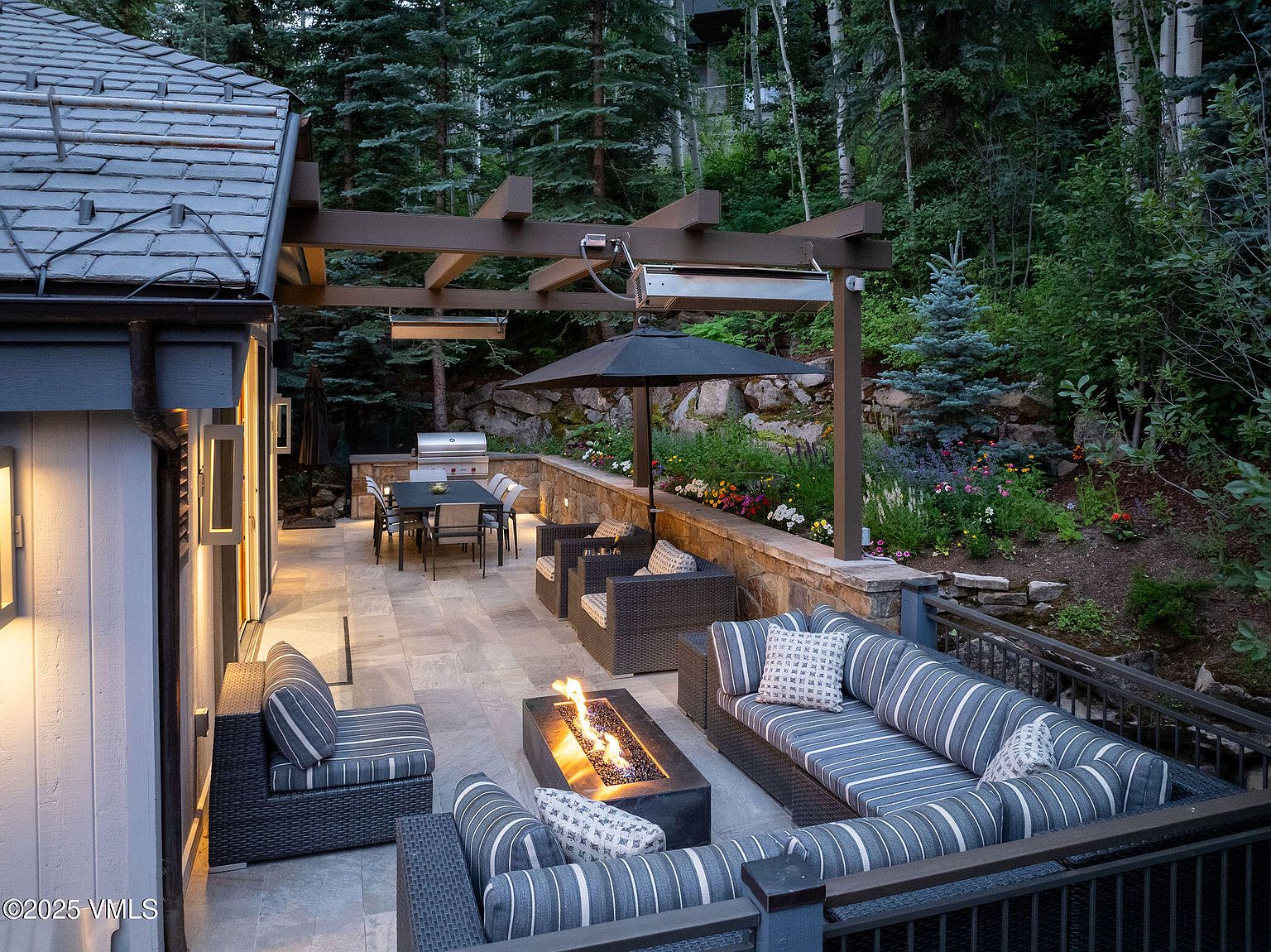 This image showcases a luxurious outdoor patio area, perfect for entertaining or relaxing. The space features comfortable seating arrangements around a modern fire pit, a dining table with a grill, and a well-maintained garden with colorful flowers. The patio is partially covered by a pergola, providing shade and adding architectural interest, while the surrounding trees create a sense of privacy and tranquility.