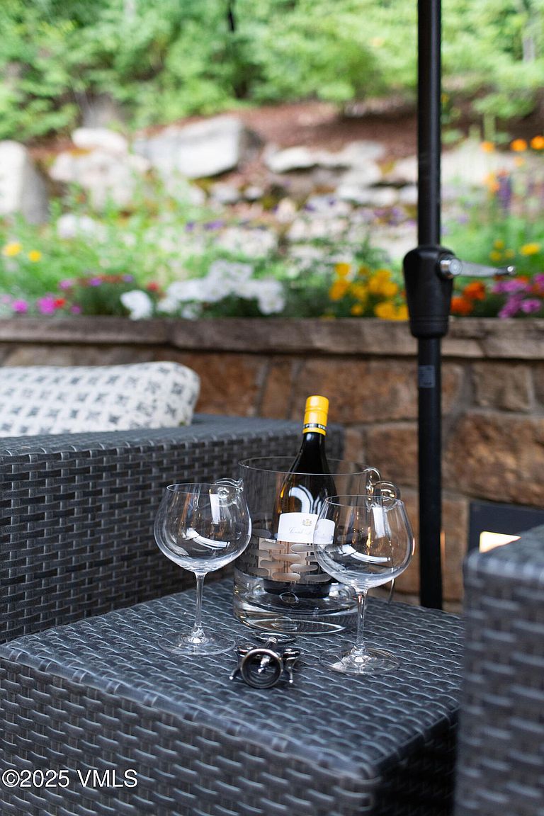 This image showcases a cozy outdoor patio setting, featuring a wicker table set with wine glasses and a bottle of wine in an ice bucket. The patio is furnished with comfortable seating, and a backdrop of lush greenery and flowers adds to the inviting atmosphere. An umbrella provides shade, enhancing the appeal of this outdoor living space.