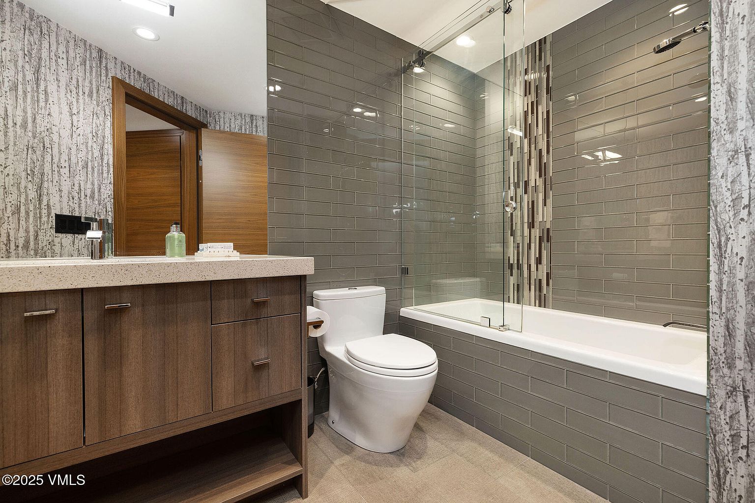 This is a modern bathroom featuring a floating vanity with a light countertop and dark wood cabinets. A white toilet sits next to the vanity, and a bathtub with a glass enclosure is tiled with gray subway tiles and a decorative vertical stripe. The overall impression is clean and contemporary.