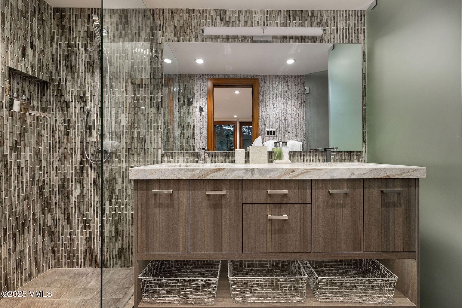 This is a primary bathroom featuring a double vanity with a light-colored countertop and wood-grain cabinets. The walls are tiled with a mosaic pattern, and there are wire baskets on the shelf below the vanity for storage. A glass-enclosed shower is visible to the left, and the overall style is modern and luxurious.