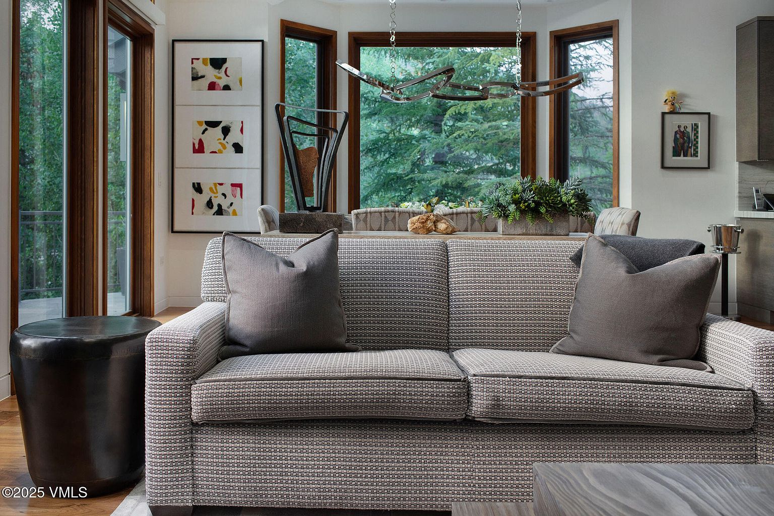 This interior shot showcases a stylish living room with a patterned sofa adorned with gray pillows, complemented by a dark metal side table. The room features large windows offering a view of lush greenery, enhancing the connection to nature. Artwork and a unique chandelier add to the sophisticated ambiance, creating a comfortable and inviting space.