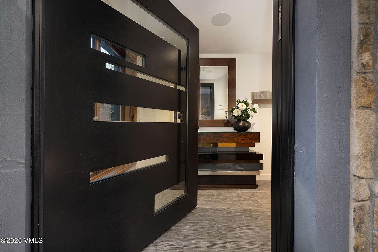This image showcases an entryway with a modern, dark wood door featuring horizontal glass panels, partially open to reveal a stylish interior. Inside, a console table with a mirror above it is adorned with a floral arrangement, set against a backdrop of neutral-toned walls and tiled flooring. The overall impression is one of contemporary elegance and inviting design.