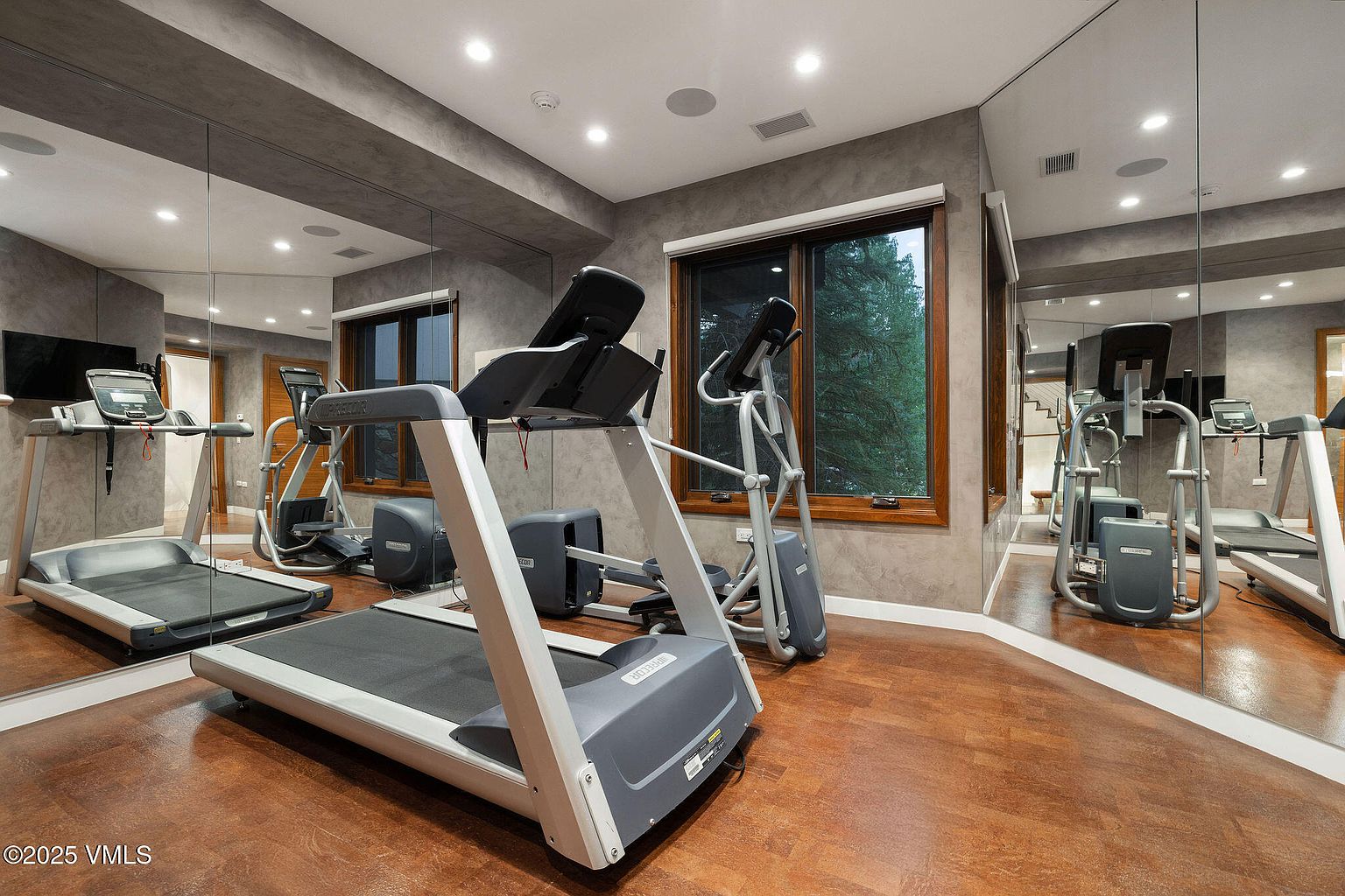 This interior shot showcases a well-equipped home gym with a treadmill and elliptical machine as the main features. The room has a modern aesthetic with gray walls, wood flooring, and large mirrors that enhance the sense of space. A window provides natural light and a view of the outdoors.