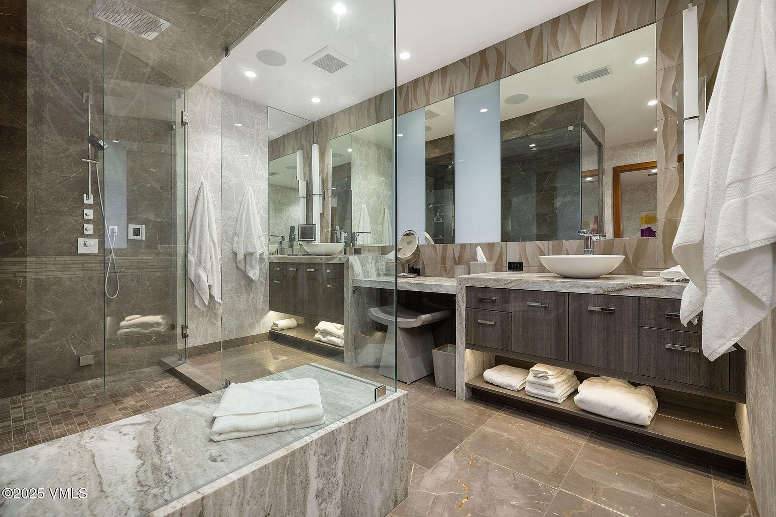 This is a luxurious primary bathroom featuring a glass-enclosed shower with multiple showerheads and a built-in bench. The double vanity has vessel sinks, modern cabinetry with under-cabinet lighting, and a large mirror. The bathroom is finished with marble-like tile flooring and walls, creating a spa-like atmosphere.