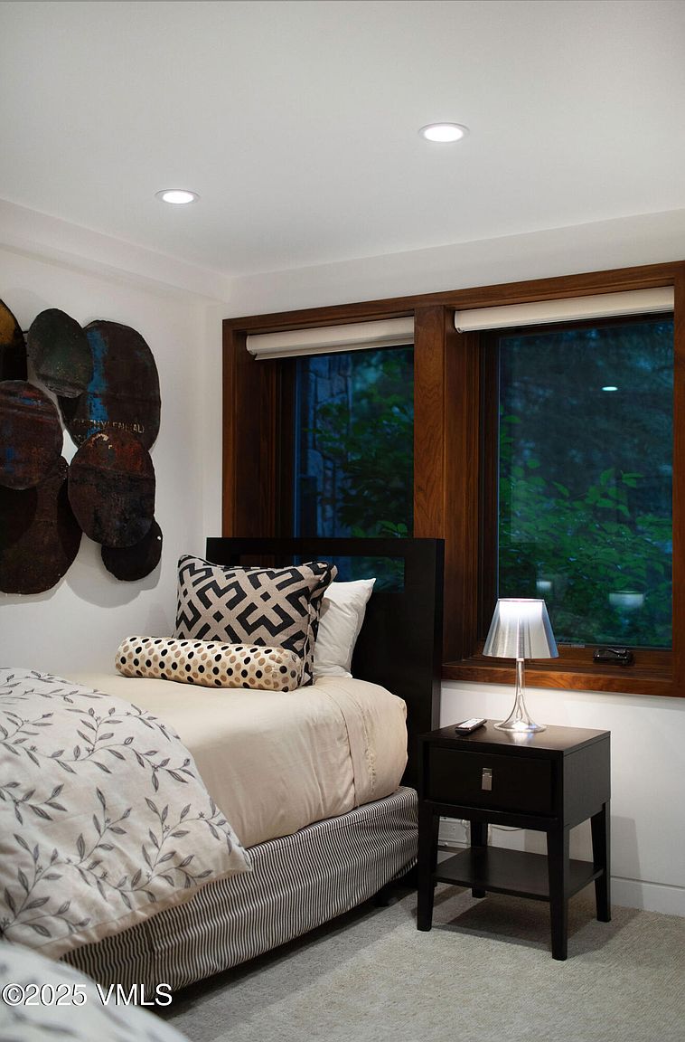 This is a cozy guest bedroom featuring a single bed with a patterned comforter and decorative pillows. A dark wood nightstand with a lamp sits beside the bed, and two windows with wood trim offer natural light. The room is decorated with modern art and has a clean, minimalist style.